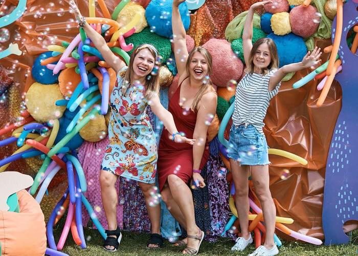 Three people in shorts and dresses stand in front of a backdrop of colorful fabrics and balloons. They smile happily at the camera with arms outstretched in excitement.