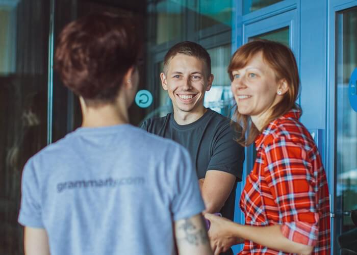 Three people smile at each other outside a Grammarly office building. The person closest to the camera has Grammarly.com written on the back of their shirt.